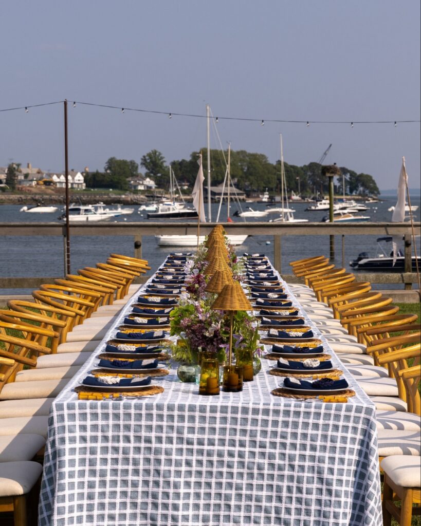 Full-service luxury wedding catering setup with long tables and wishbone chairs overlooking a waterfront in the Hudson Valley.